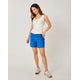 A woman stands against a plain background, wearing a white tank top, Carve Oahu 6" Shorts in bright blue stretch cotton corduroy, and sneakers—an effortless addition to your vacation wardrobe.