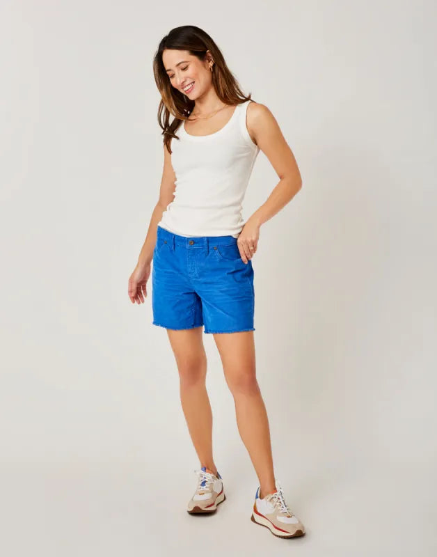 A woman stands against a plain background, wearing a white tank top, Carve Oahu 6" Shorts in bright blue stretch cotton corduroy, and sneakers—an effortless addition to your vacation wardrobe.