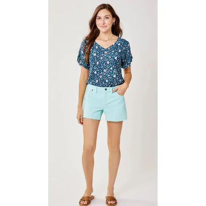 Woman standing against a plain background wears Carve's Oahu 6" Short in light blue, a blue floral blouse, and brown sandals with hands in pockets—an ideal addition to your vacation wardrobe.