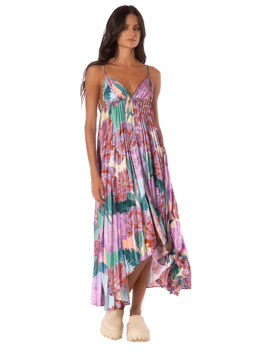 A woman models the Maaji Parrot Dress—a colorful, floral, sleeveless maxi with a handkerchief hem—paired with beige platform shoes, set against a white background.