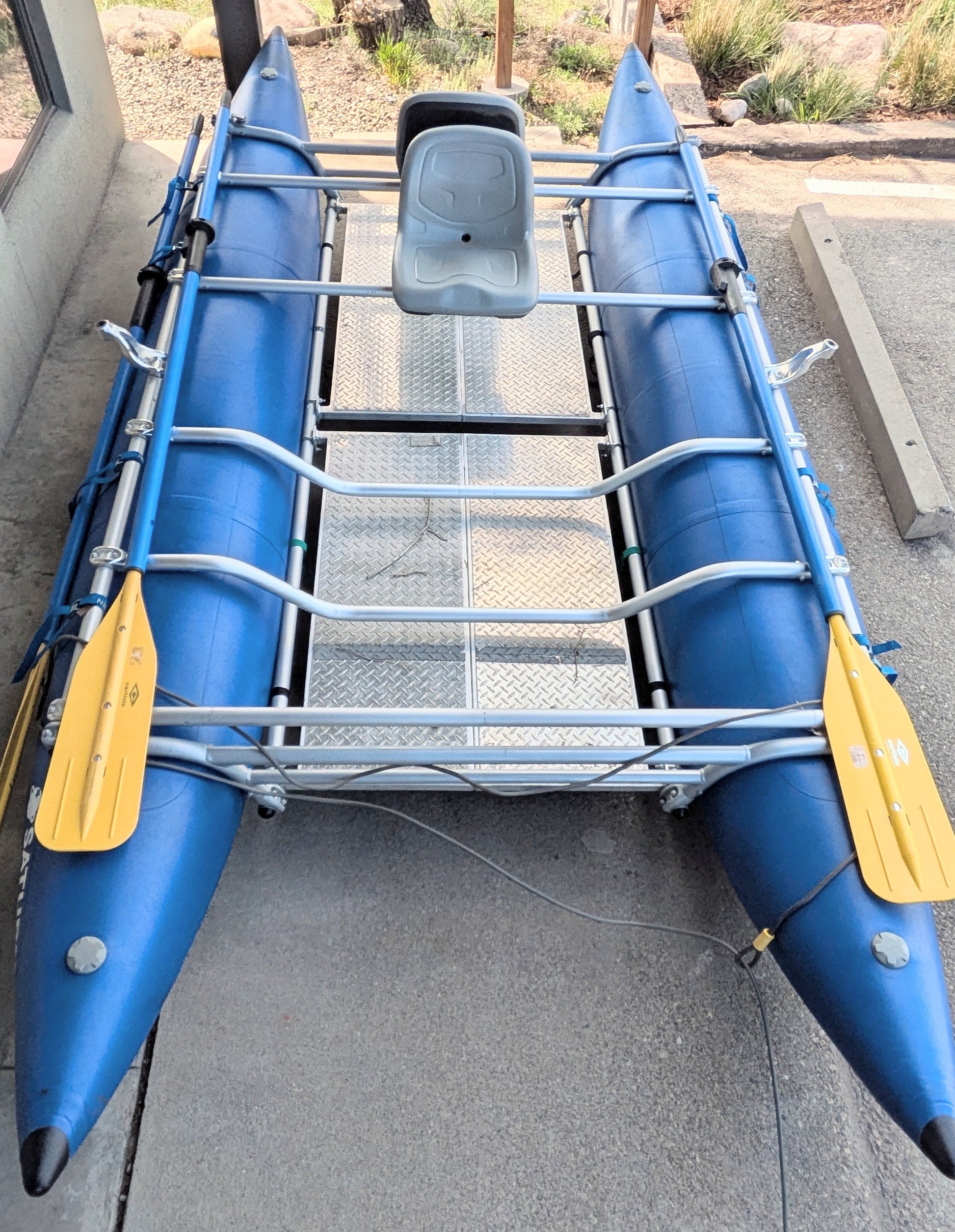 A 16' Cataraft Saturn by 4Corners Riversports, featuring a metal frame, single grey seat, and two yellow paddles, sits on concrete—ideal for fishing or whitewater trips.