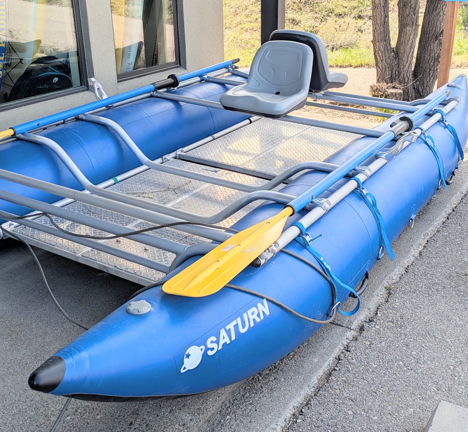 A 16' Cataraft Saturn by 4Corners Riversports, featuring a blue inflatable hull, metal frame, oars, and a gray plastic seat for fishing or whitewater use, sits on pavement next to a building.