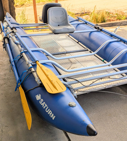 The 16' Cataraft Saturn by 4Corners Riversports, a blue inflatable raft with yellow paddles attached to the side, is set on a concrete area near grass and a tree, ideal for fishing or whitewater use.