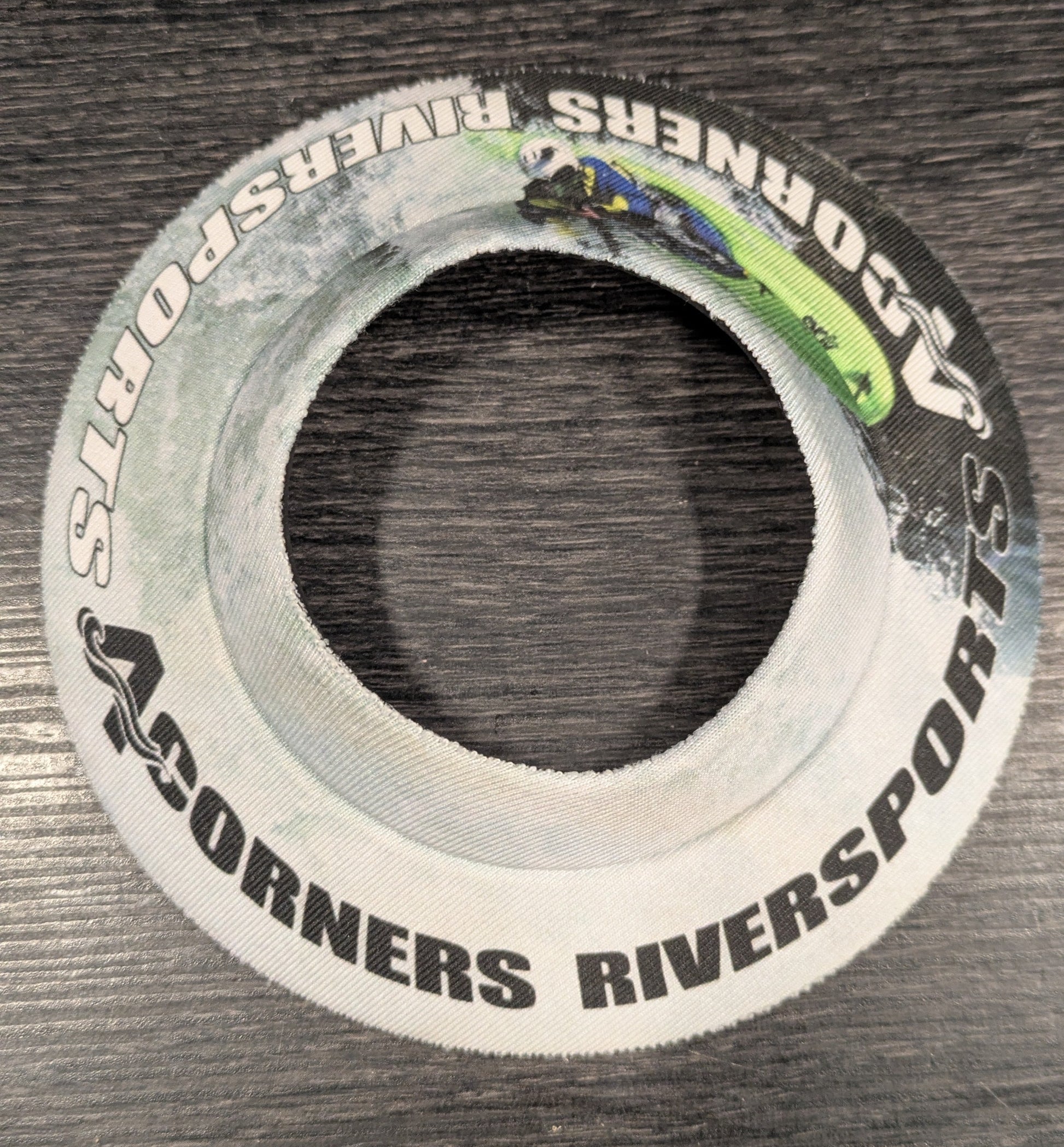The Lil' Sucker Boozie by 4CRS is a round white fabric band with "4Corners Riversports" text and a kayaker image, designed to securely hold your beer. Shown on a dark wood surface.