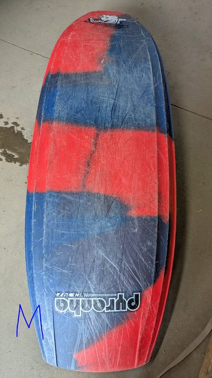 A worn surfboard with red and blue stripes, scratches, and a “Phantom” logo lies on concrete near a blue "M"—much like the Pyranha Demo Helectron used kayak from the demo fleet.