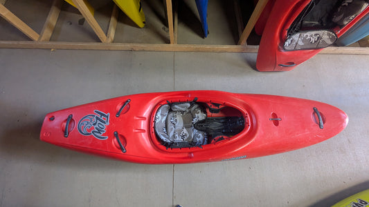 A Jackson Kayak Demo Flow SM in red with black and white interior padding is on a concrete floor, while other kayaks are stored vertically in the background.