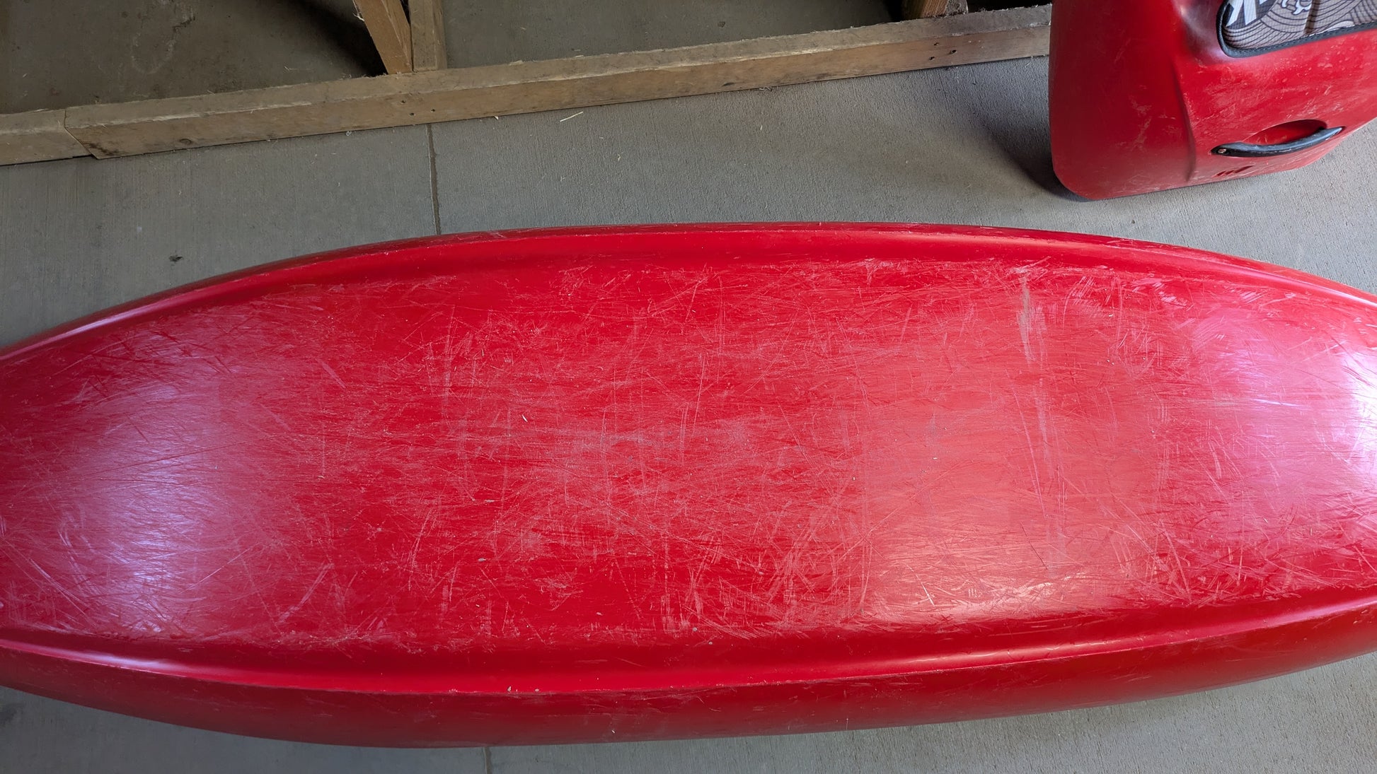 A used red Jackson Kayak Demo Flow SM with visible scratches and wear is shown from above, resting on a concrete floor next to a wooden frame.