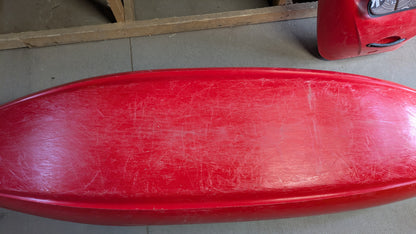 A used red Jackson Kayak Demo Flow SM with visible scratches and wear is shown from above, resting on a concrete floor next to a wooden frame.
