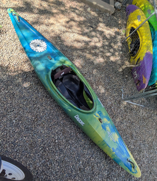 A barely used Dagger Sawblade Outburst kayak from 4Corners Riversports, featuring green and blue colors, sits on gravel near a fence with another yellow and purple kayak partially visible in the background.