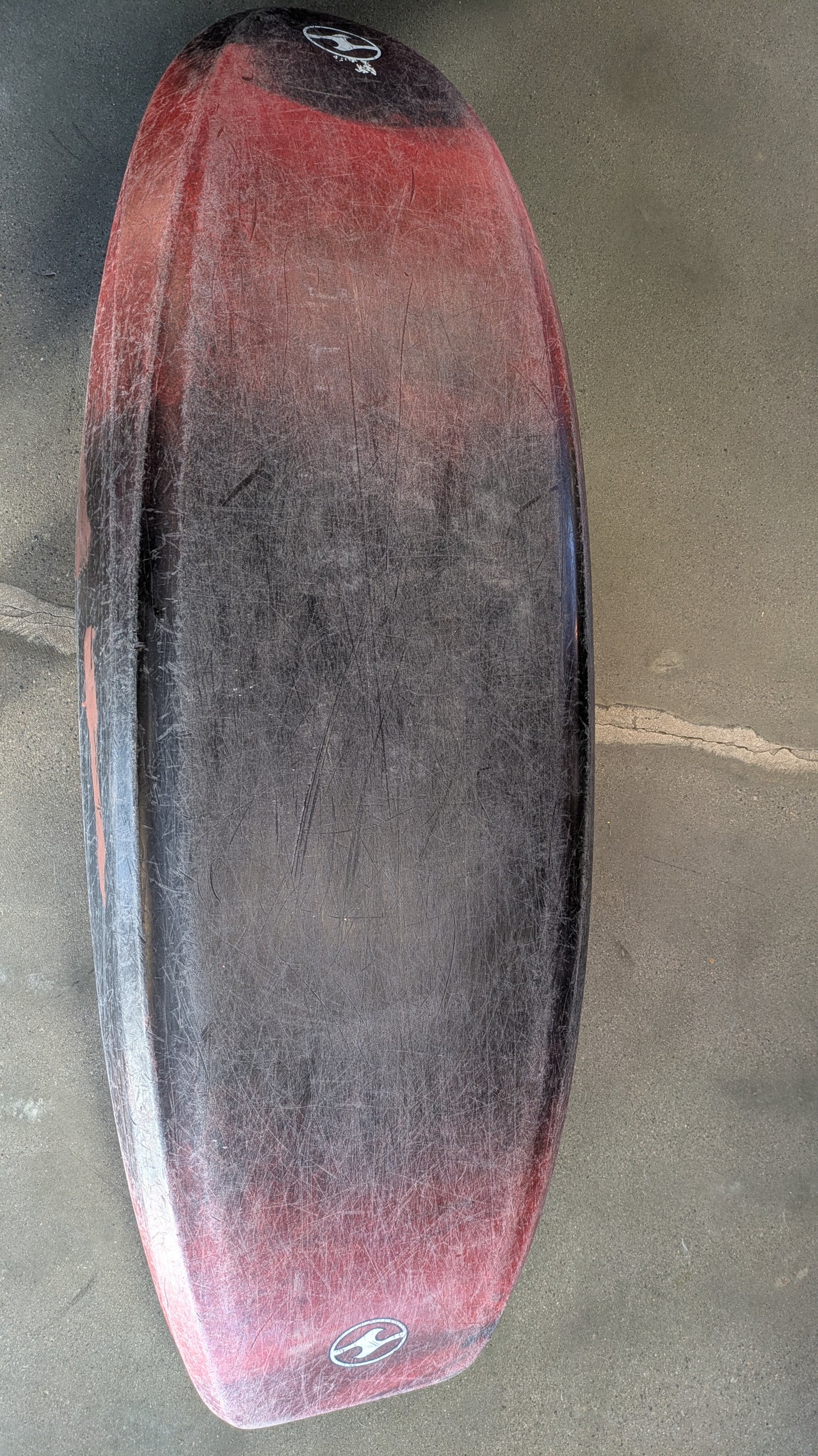 A red and black bodyboard with visible scratches lies on a concrete surface, recalling the well-worn look of a 4CRS Consignment Rockstar 4.0 kayak.