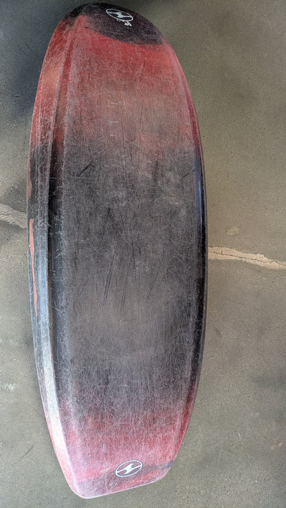 A red and black bodyboard with visible scratches lies on a concrete surface, recalling the well-worn look of a 4CRS Consignment Rockstar 4.0 kayak.