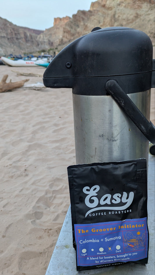 A Groover Initiator Coffee bag from 4Corners Riversports and a metal coffee thermos rest on a sandy campsite table, ideal for enjoying fresh coffee by the river and cliffs.