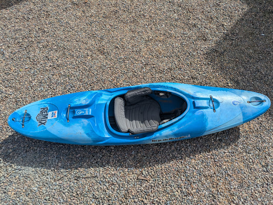 A blue Consignment LiquidLogic Remix 69 beginner kayak by LiquidLogic with a padded gray seat rests on small gravel stones.
