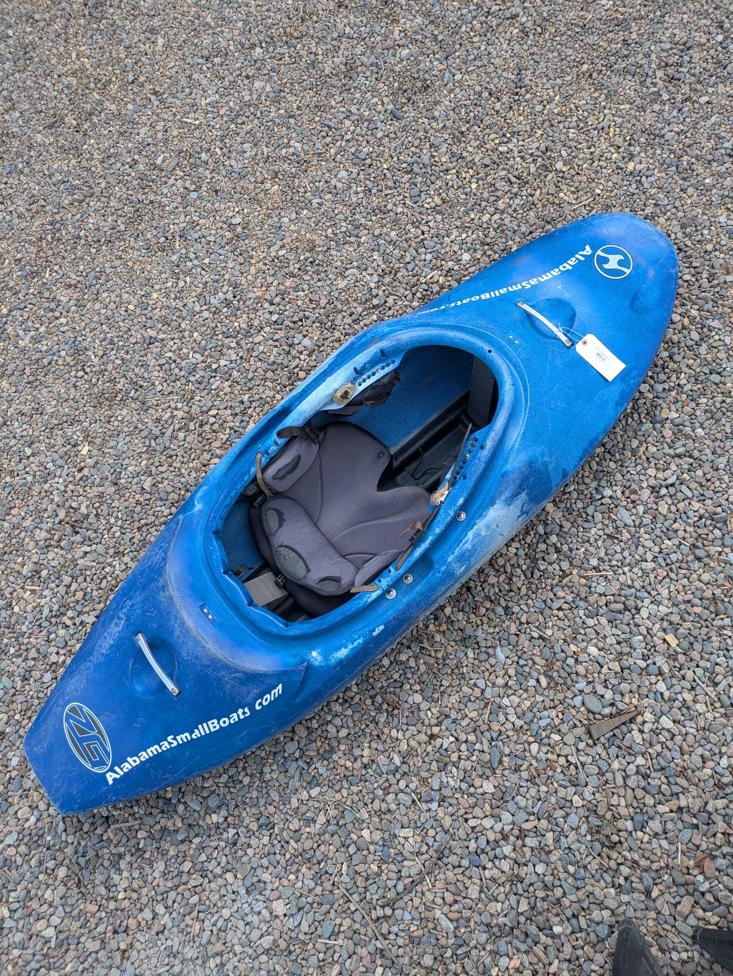 A blue Consignment Wavesport ZG kayak by Wavesport sits on gravel, featuring a padded seat and "AlabamaSmallBoats.com" printed on the side.