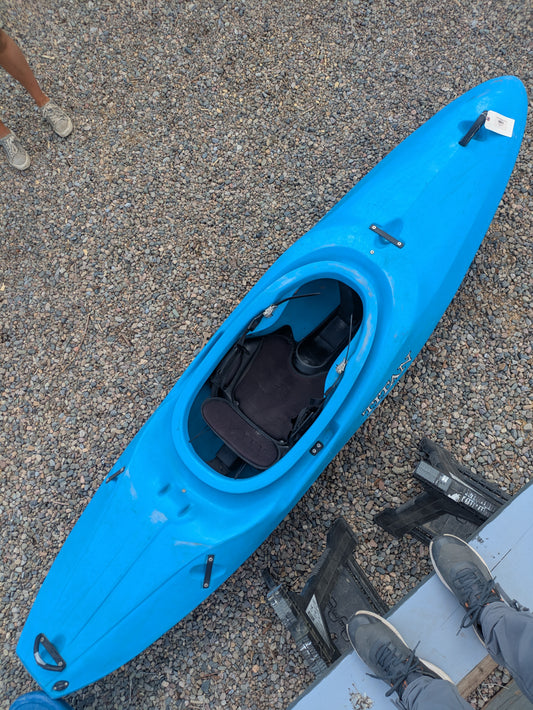 A Consignment Titan Rival kayak by Titan Kayaks in bright blue rests on a gravel surface, viewed from above, with three people’s feet partially visible around it.