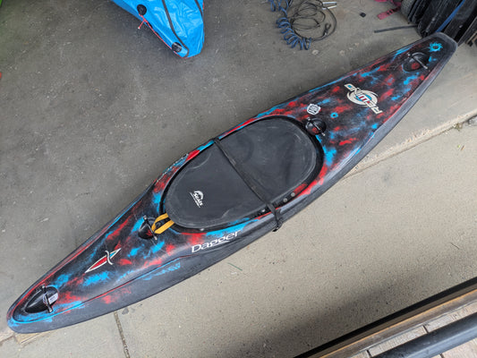 A 4Corners Riversports Consignment Rewind LG Cosmo kayak, featuring Contour Ergo outfitting and a closed cockpit, is on concrete near coiled cables and another blue kayak. The kayak has black, blue, and red colors.