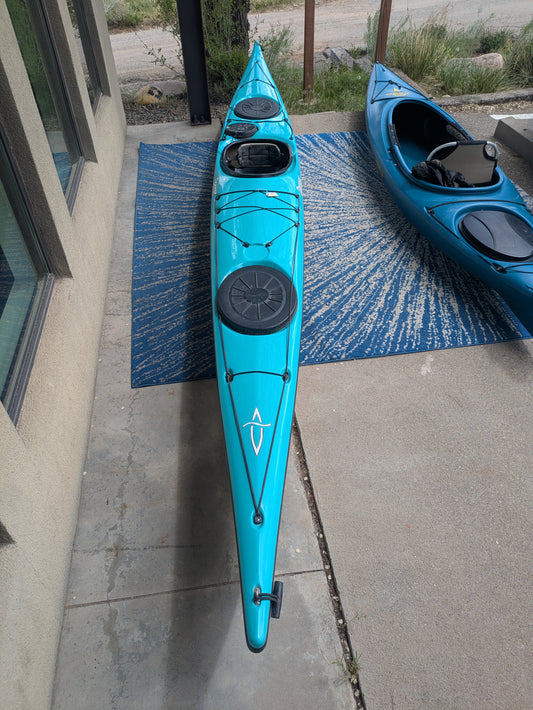 A teal Dagger Sitka Touring/Sea Kayak by Dagger, featuring black straps, storage hatches, and an integrated rudder, sits on a blue-patterned rug beside a building with another kayak partially visible nearby.