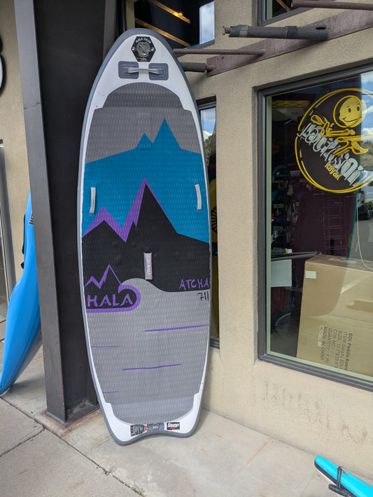 The Hala Gear Atcha 711 inflatable SUP, featuring innovative Stompbox Technology and a mountain graphic, leans against an outdoor shop window.