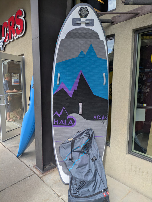 The Hala Gear Hala Atcha 711 inflatable SUP, designed for river surfing with Stompbox Technology and mountain graphics, stands against a storefront beside its gray carrying bag.