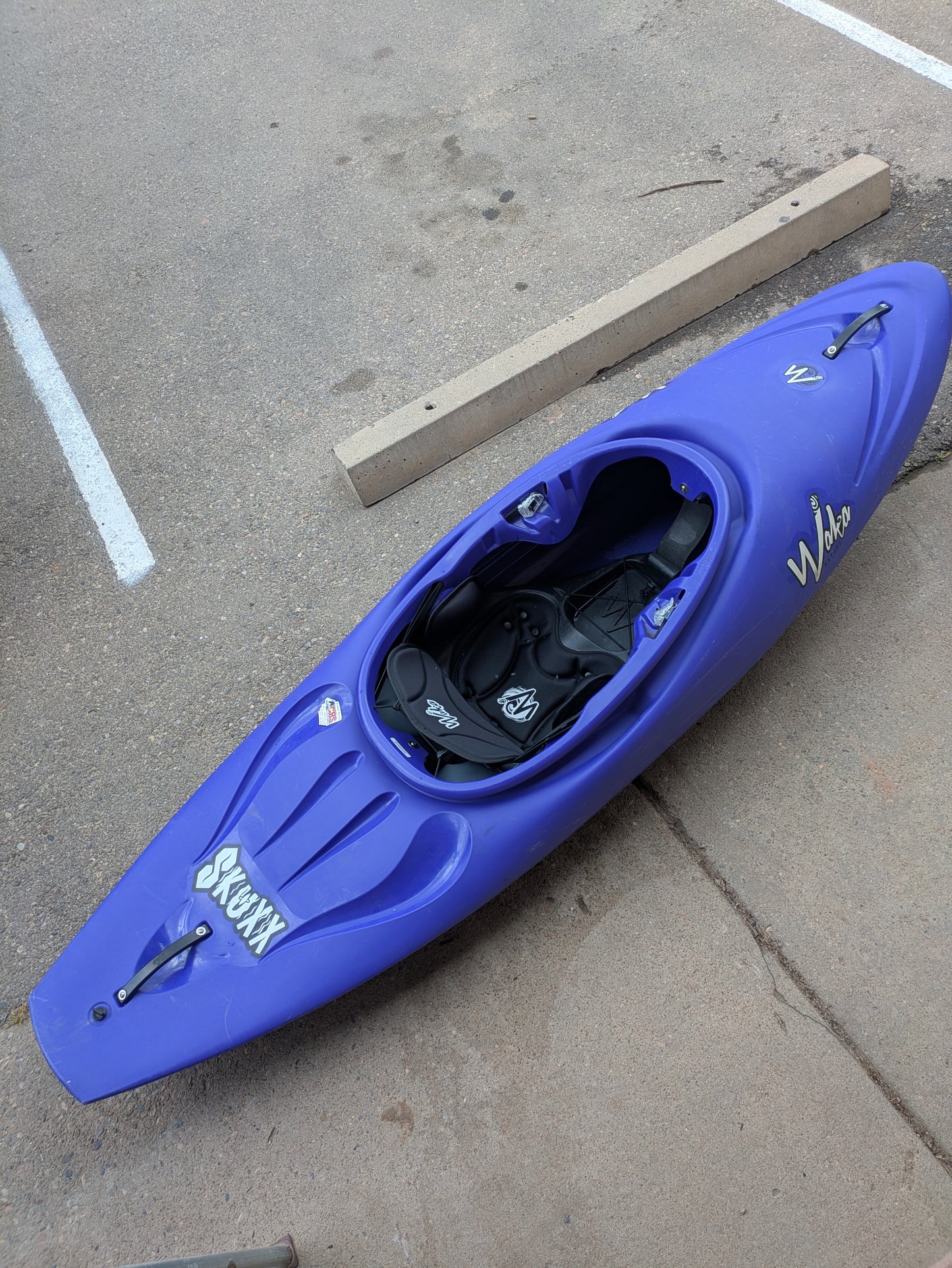 A blue river running kayak labeled "Demo Skuxx" and branded "Waka Kayaks" sits on a concrete surface near parking lines and a parking curb.