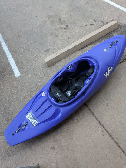 A blue river running kayak labeled "Demo Skuxx" and branded "Waka Kayaks" sits on a concrete surface near parking lines and a parking curb.