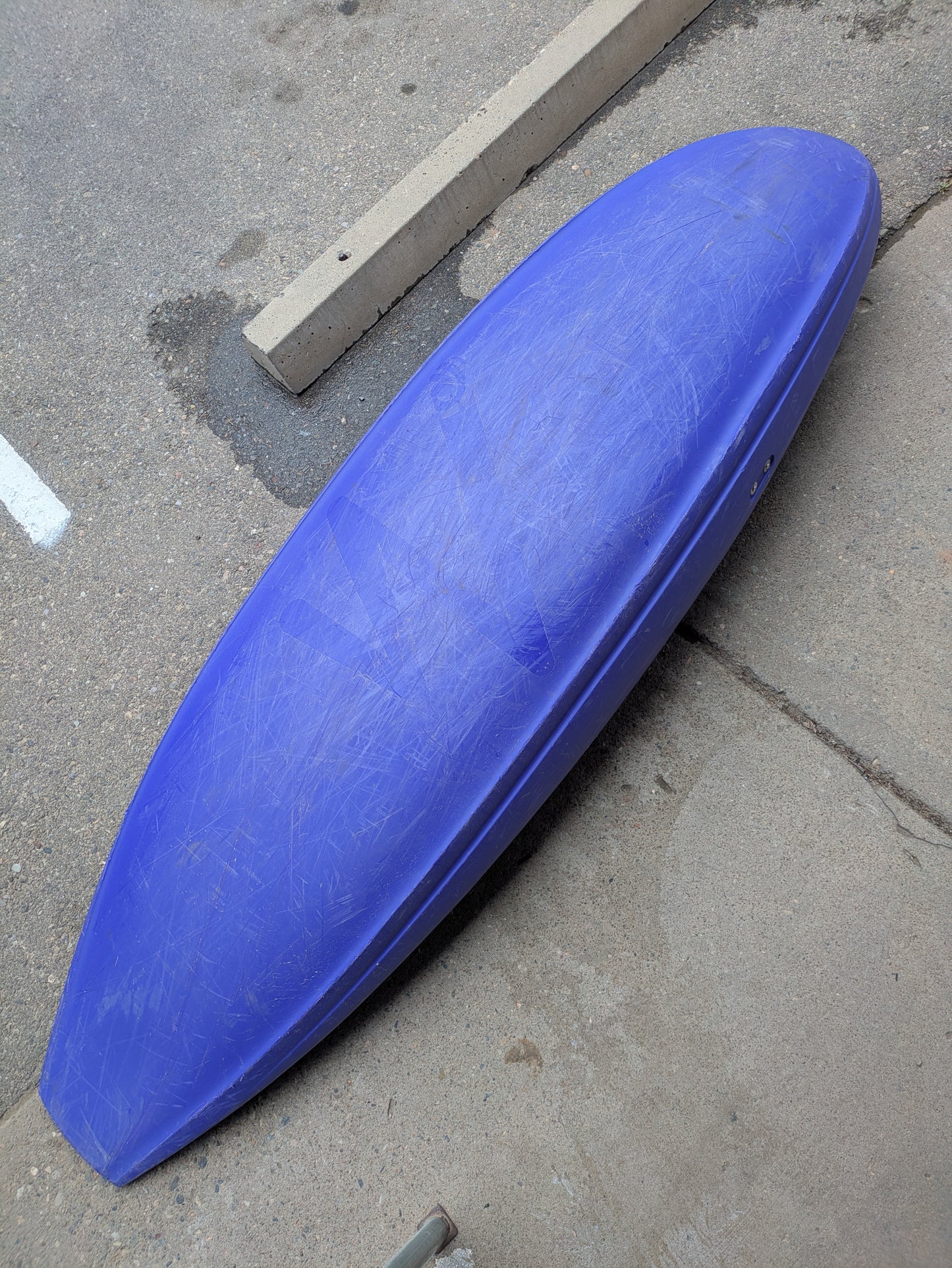 A blue Waka Kayaks Demo Skuxx kayak is lying upside down on a concrete surface next to a parking curb.