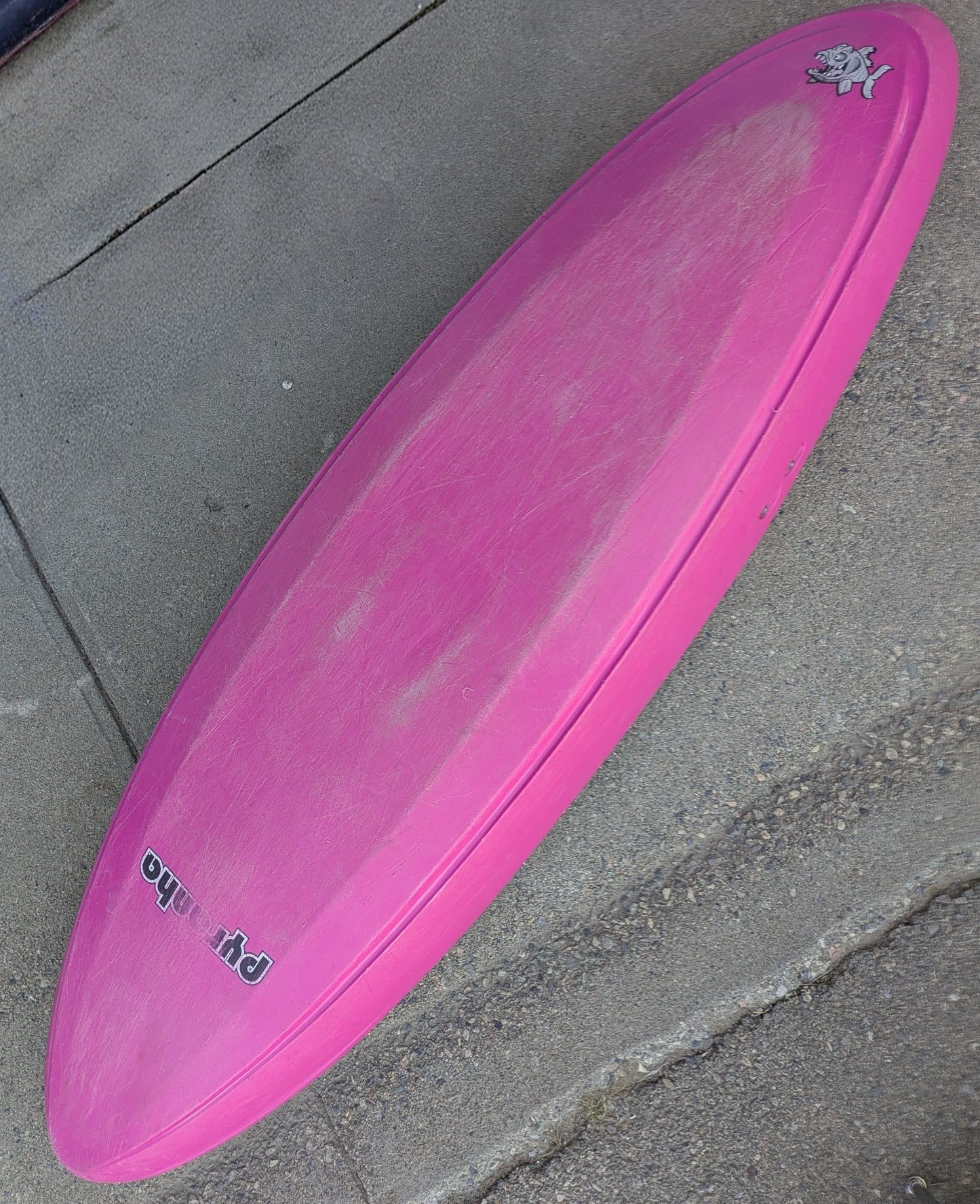 A Consignment Pyranha Burn 1 - Small Pink kayak from 4Corners Riversports lies upside down on concrete, showing signs of use and featuring a small cartoon graphic near the nose.