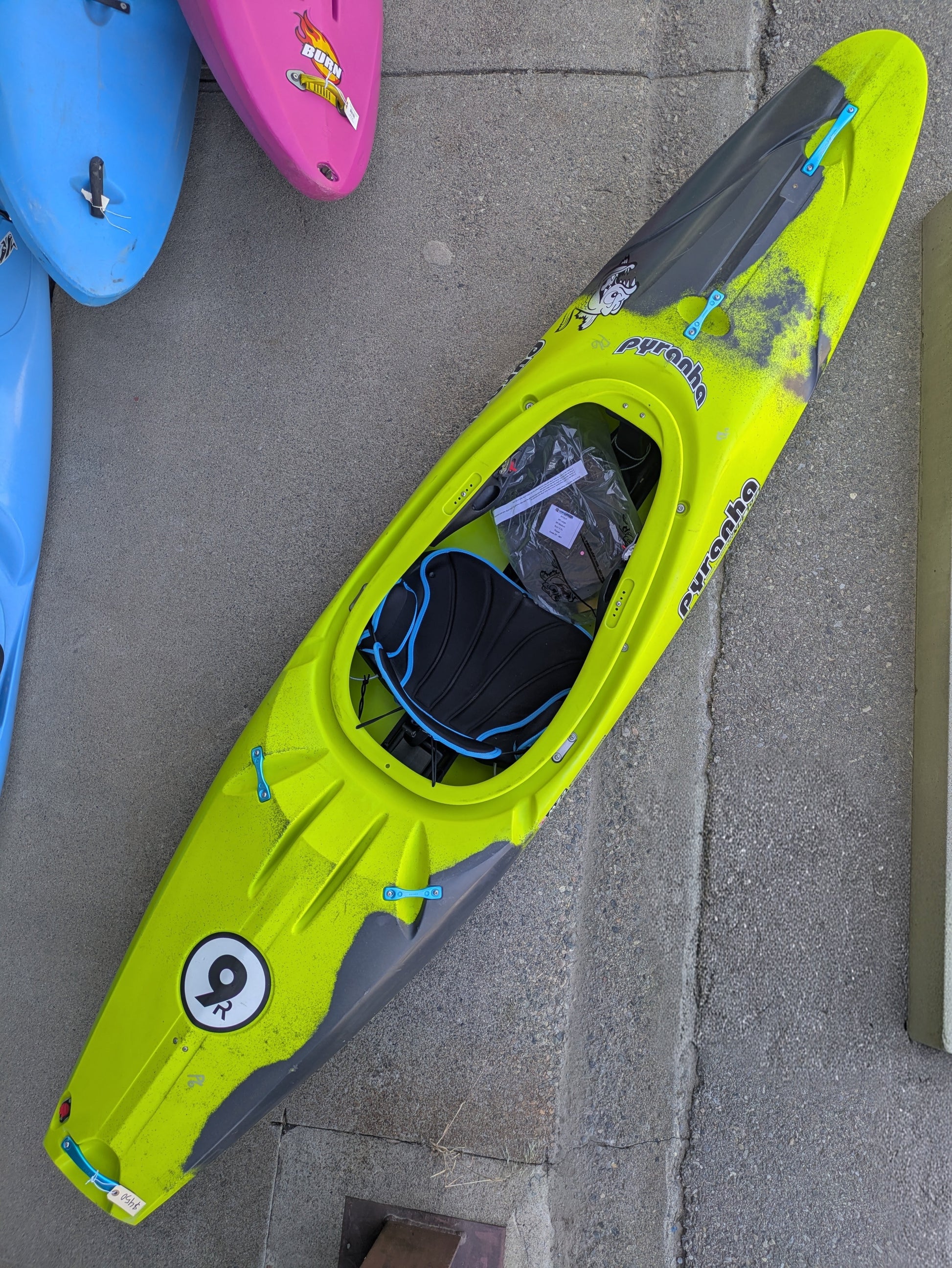 A Consignment Pyranha 9R Large kayak by 4Corners Riversports, featuring lime green with black and blue accents, rests on concrete among other kayaks.