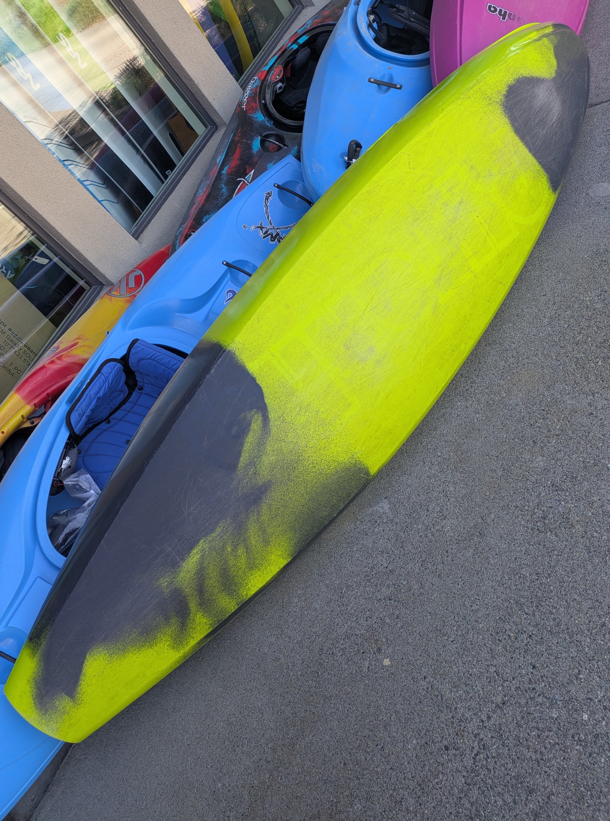 A Consignment Pyranha 9r Large kayak from 4Corners Riversports, in yellow and black, lies upside down on a concrete surface beside several other colorful kayaks.