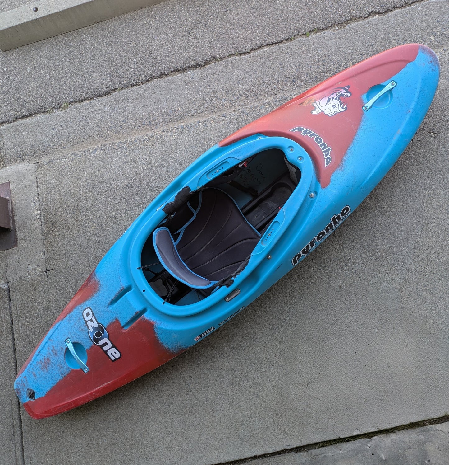 A Consignment Medium Ozone kayak from 4Corners Riversports, featuring a red and blue design, sits on a concrete surface shown from above with its seat and interior visible.