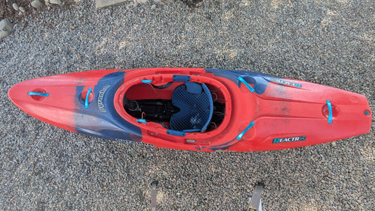 Top view of a used Pyranha Demo ReactR LG kayak in red and blue on gravel. The kayak has a blue seat and visible signs of use, indicating it was previously a demo boat.