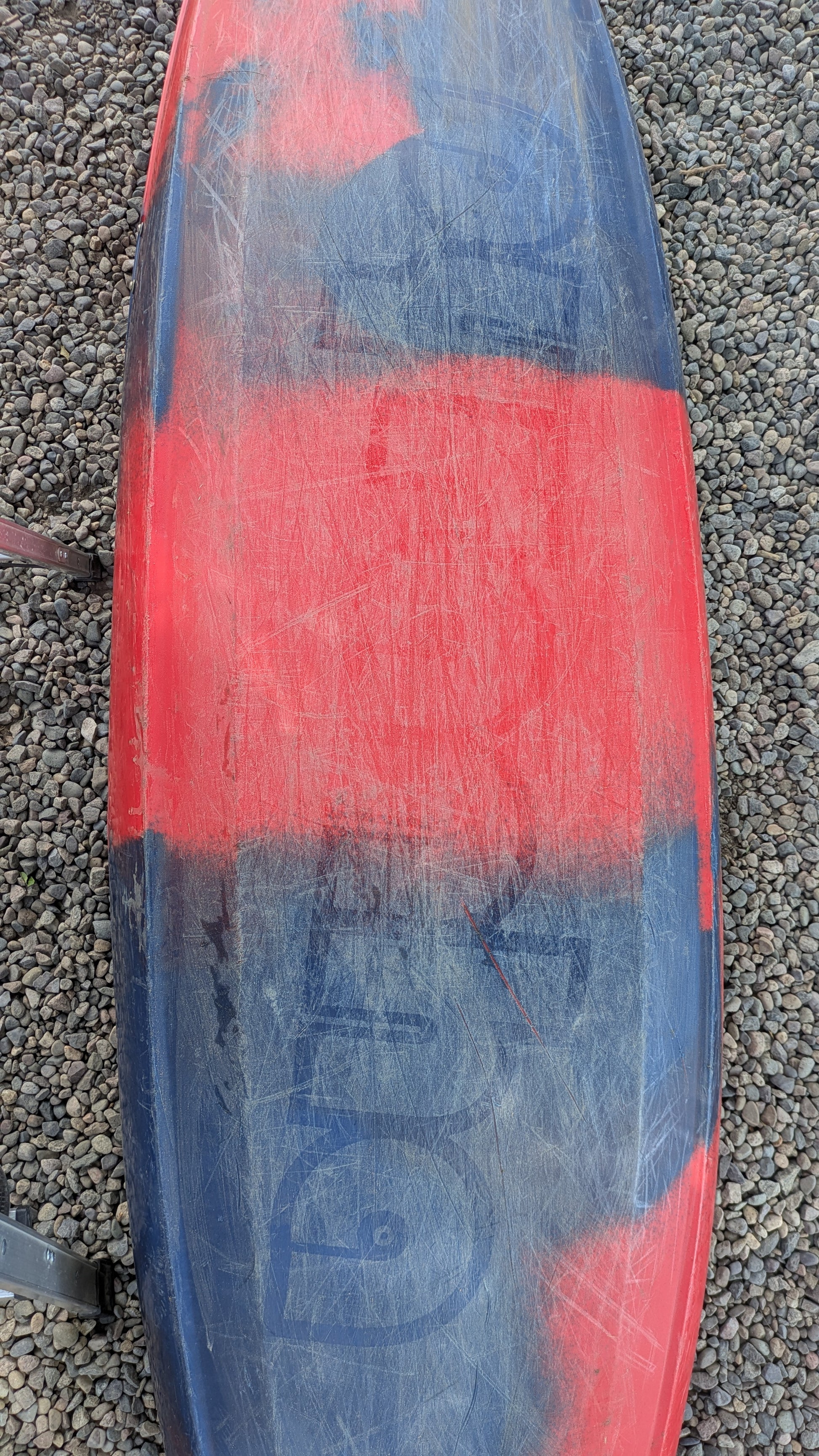 A scratched and worn red and black Pyranha Demo ReactR LG kayak rests on small gravel stones.