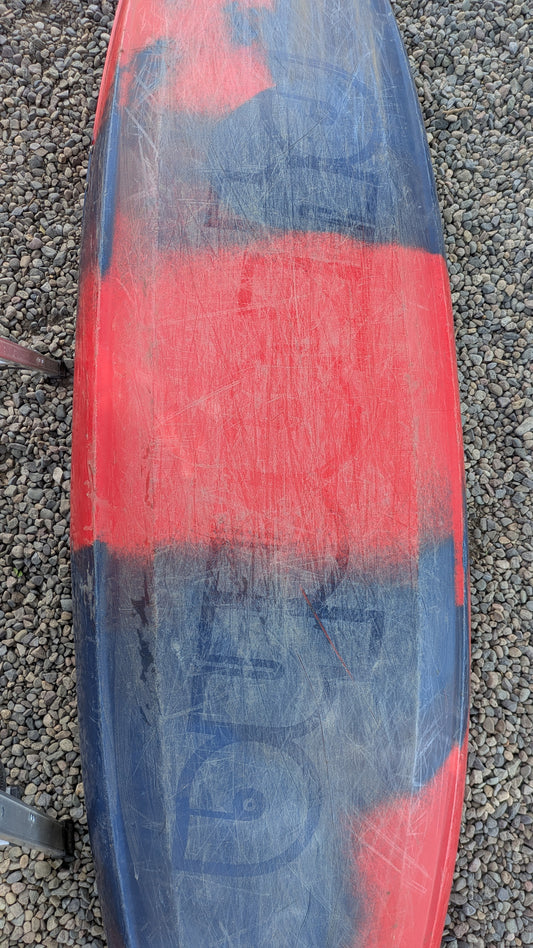 A scratched and worn red and black Pyranha Demo ReactR LG kayak rests on small gravel stones.