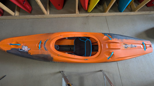 A Pyranha Demo Scorch LG kayak in orange with blue handles and a graphic sticker is seen from above, resting on a concrete floor near a storage rack holding other kayaks.