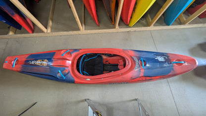 A Pyranha Demo Scorch X kayak in red and blue stands upright on a concrete floor, with other colorful kayaks stored vertically in the background.