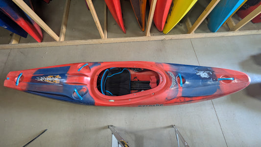 A Pyranha Demo Scorch X kayak in red and blue stands upright on a concrete floor, with other colorful kayaks stored vertically in the background.