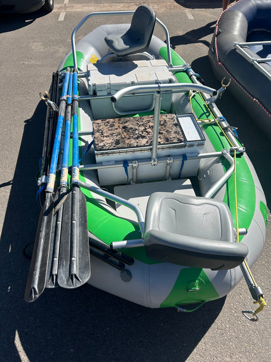 A green and gray 4CRS Rental Raft - Fishing Package with two seats, side oars, and a central storage box, ideal for multi-day trips, is set on a paved surface.