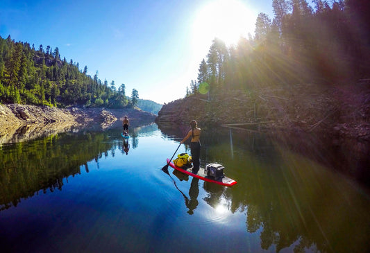 Two people paddleboard on a calm, tree-lined lake under blue skies, gear secured—ideal for anyone seeking a peaceful day with 4CRS Rental SUP inflatable boards.
