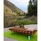 A vibrant FunLuvin' Cuddle Buddy from FunLuvin' Fleecewear, featuring a cozy fleece and cotton cover, is set up beside a river, surrounded by hills and trees. A backpack and water bottle are placed on the green mat beneath it, completing the ideal camping sleep gear setup.