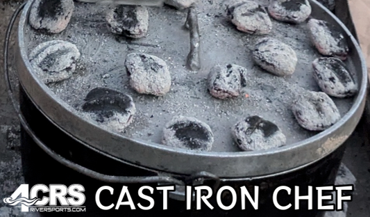 A 4CRS Cast Iron Chef pot with a lid, covered in round charcoal briquettes, ideal for riverside outdoor cooking.