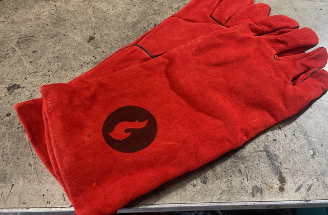 A pair of red protective gloves with a flame icon rests on scratched metal beside the SWOK Discada Combo by LavaBox—an essential accessory for safe campfire cooking and handling hot outdoor cookware.