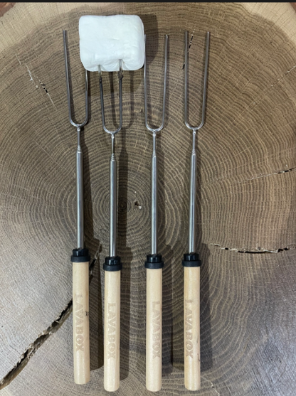 Four SWOK Discada Combo roasting forks by LavaBox, each with wooden handles, are arranged on a tree stump for campfire cooking; the second fork from the left holds a marshmallow ready to be toasted.