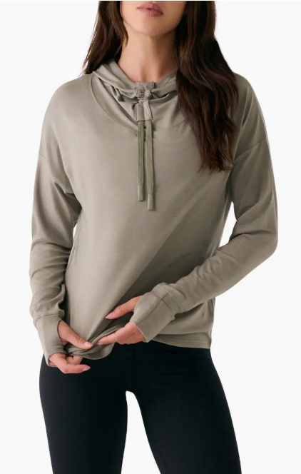 A woman wears the Lole Dynamic Hoodie in light olive green—featuring breathable knit fabric, a drawstring neck, and thumb holes—paired with black leggings.