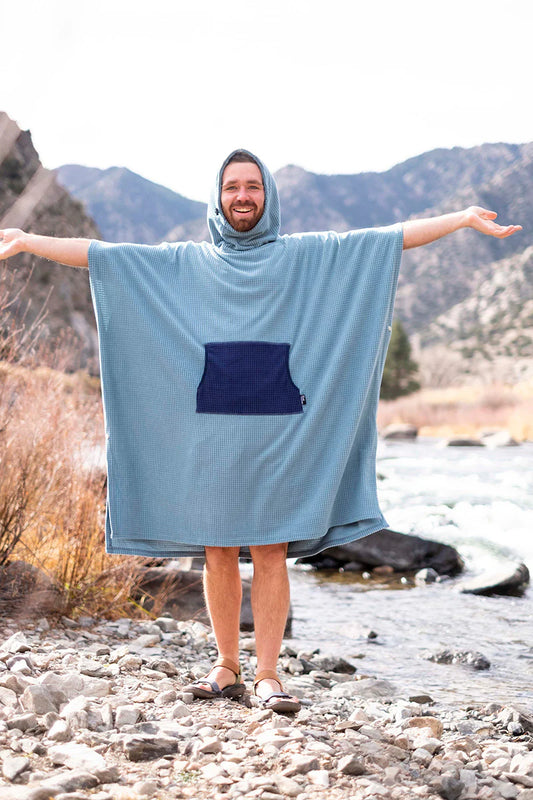 A man stands on rocky ground by a river, smiling with arms outstretched, wearing the River Station Gear Fleece Poncho Changing Robe in light blue with a front pocket and sandals. Mountains rise in the background.