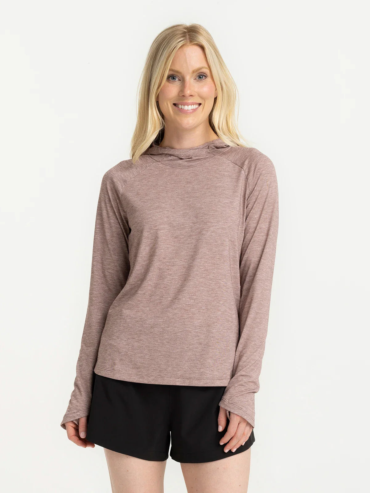 A woman with long blonde hair wears a mauve Free Fly Elevate Hoodie W's featuring drirelease tech and black shorts, standing and smiling against a plain white background.