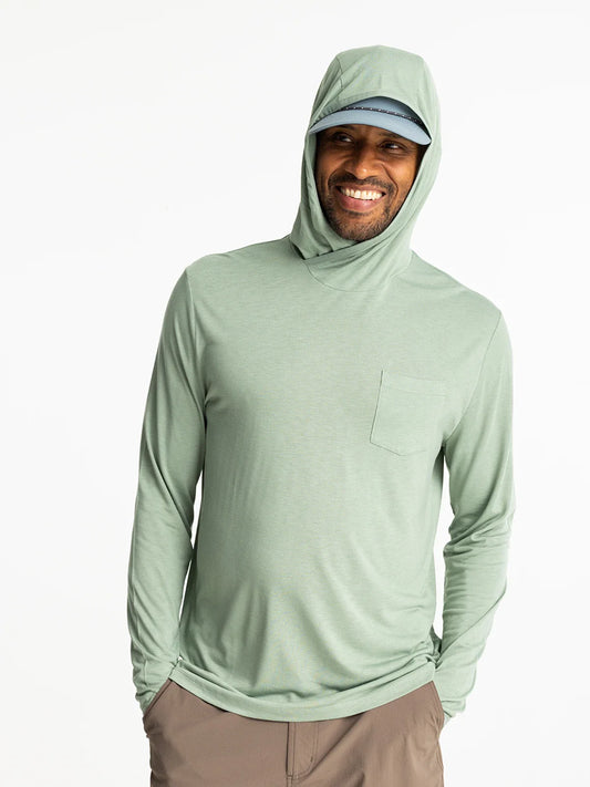 A man in a light green Free Fly Bamboo Lightweight Hoodie M's, a light blue cap, and brown pants stands smiling against a white background—ideal for fishing or sun protection.