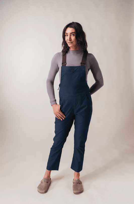 A person stands against a light background wearing the Allover Lookout Overalls 2.0 W's in blue with adjustable straps, paired with a grey long-sleeve shirt and beige slip-on shoes.