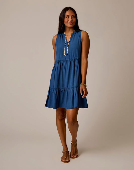 A woman stands in a studio against a beige background wearing the Carve Nellie Dress, an elegant sleeveless blue tiered piece with side pockets, paired with tan sandals and a long necklace.