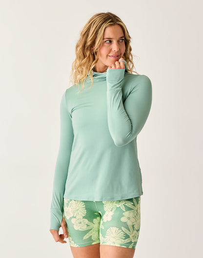 A woman wears the Carve Alani Sunshirt in light green with green floral shorts, standing against a plain background and smiling to the side.