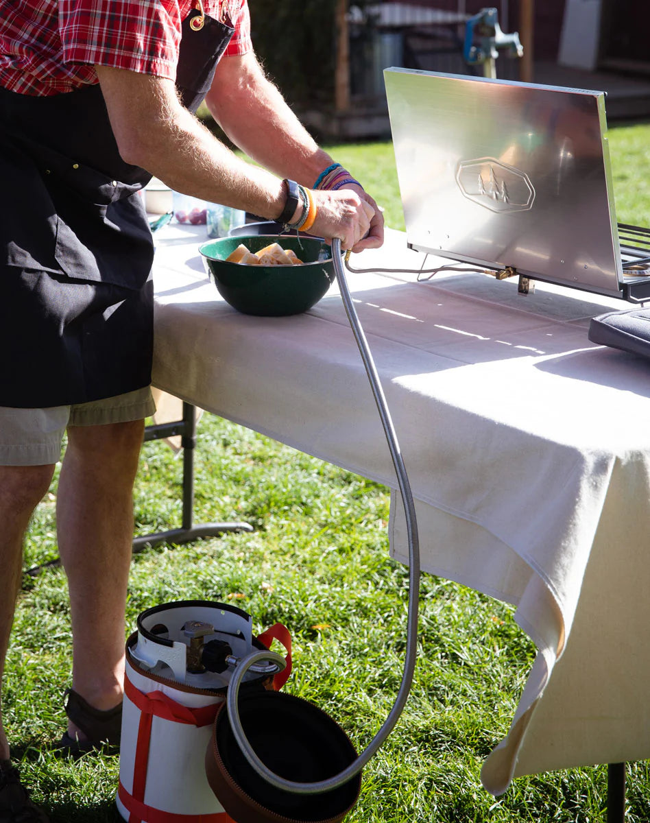 Someone is connecting a propane tank to a portable grill on an outdoor table using the GSI Outdoors Propane Adapter Hose, which is sturdy and made of stainless steel. A bowl sits nearby for grilling prep.